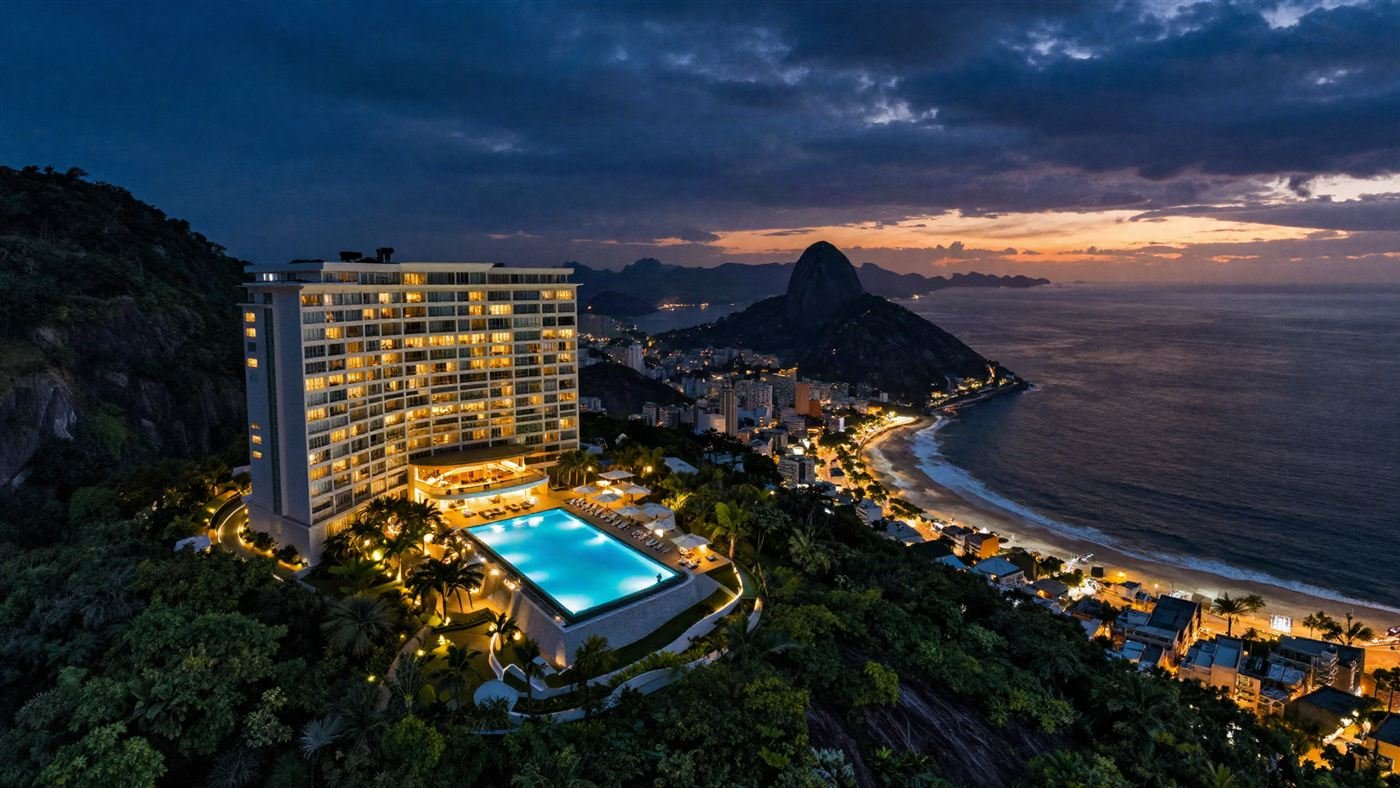 Vista aérea de um hotel com piscina iluminada na encosta, com o Pão de Açúcar ao fundo, na hora azul